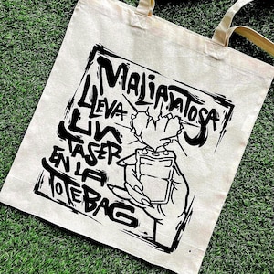 Puede incluir: Bolso tote beige con un diseño gráfico negro. El diseño presenta las palabras "Maliciosa" y "Lleva un laser en la tote bag" en un estilo audaz y manuscrito, junto con una mano sosteniendo un láser. El bolso tiene dos asas beige.