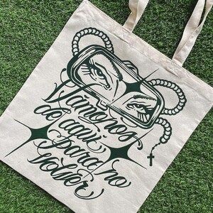 May include: Beige canvas tote bag with dark green graphic design. The design includes a stylized eye image within a rectangular frame, a rosary, and text in a cursive font. The bag has two beige handles.