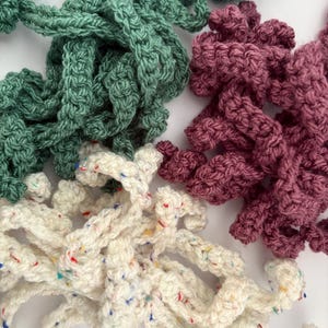 Può includere: Primo piano di quattro fili di filato all'uncinetto nei toni del verde, bordeaux e bianco screziato. Il filato è spesso e strutturato, con un aspetto fatto a mano. Il filato bianco ha macchie colorate.