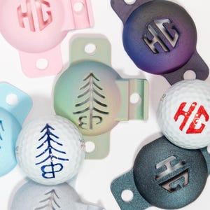 May include: A selection of golf ball markers and golf balls. The markers are in various colours, including pink, blue, green, purple, and grey, and feature the initials "HG" or a tree design. The golf balls have the same designs in red and blue.