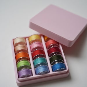 Könnte beinhalten: Eine offene, rosa Nähkiste mit einer Vielzahl farbiger Garnrollen. Die Rollen sind in Reihen angeordnet und zeigen ein Spektrum an Farben wie Rosa, Gelb, Orange, Grün und Blau. Die Box steht auf einer weißen Oberfläche.