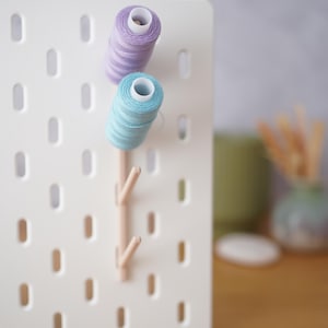 Può includere: Un pannello forato bianco con fessure ovali verticali presenta due pioli in legno chiaro inseriti nel pannello. Una bobina di filo color lavanda è posizionata sul piolo superiore e una bobina di filo azzurro è sul piolo sottostante.