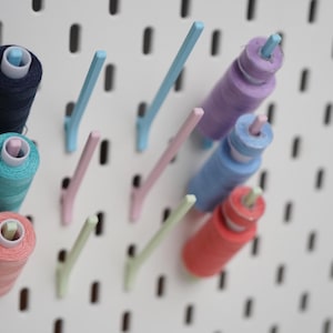 Könnte beinhalten: Eine weiße Lochplatte mit drei vertikalen Reihen von Nähgarnrollen in Marineblau, Türkis, Koralle, Lavendel, Blau und Rot. Die Garnrollen stecken auf hellblauen, rosafarbenen und hellgrünen Kunststoffstiften, die aus der Platte herausragen.