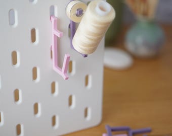 Könnte beinhalten: Eine weiße Lochplatte mit vertikalen Schlitzen und angebrachten violetten und rosafarbenen Kunststoffhaken. Ein violetter Haken hält eine Spule mit beigem Garn. Weitere violette und rosafarbene Kunststoffhaken liegen im Vordergrund.