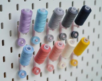 Könnte beinhalten: Eine weiße Lochwand als Wandorganizer mit mehreren Nähgarnrollen in verschiedenen Farben wie Lavendel, Hellblau, Koralle, Rosa, Creme, Gelb, Grau und Dunkelblau, die auf kleinen, in einem Raster angeordneten rosa Kunststoffstiften stecken.