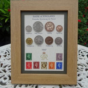 May include: A light wood frame containing a 1947 Bank of England display with eight vintage coins labeled Sixpence, Half Crown, Penny, Farthing, Threepence, Florin, Halfpenny, and Shilling, plus seven colourful postage stamps and a gold GvR emblem.