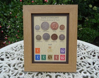 May include: A light oak-colored wooden frame containing a collection of 1956 British coins and postage stamps. The display features the text Bank of England, Pounds, Shillings & Pence, and the royal cypher ER with a crown symbol above the year 1956.