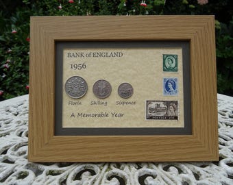 Puede incluir: Un marco de madera clara que contiene una muestra conmemorativa de 1956 del Banco de Inglaterra. Incluye tres monedas de plata etiquetadas como Florin, Shilling y Sixpence, junto a tres sellos postales con la reina Isabel II y un castillo.
