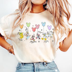 May include: Cream-colored t-shirt featuring a colorful graphic of Disney characters holding Mickey Mouse-shaped balloons. The characters include Pluto, Goofy, Mickey, Minnie, Donald, and Daisy Duck. The shirt has a relaxed fit.