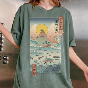 May include: Sage green t-shirt featuring a Japanese-inspired graphic. The design depicts a boat on a wave, a rising sun, and Japanese text. The shirt has short sleeves and a relaxed fit.