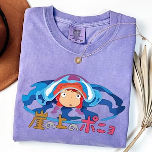 May include: A lavender Comfort Colors t-shirt featuring a cartoon character with red hair and a blue and white head covering. Japanese text is printed below the character. A gold necklace with a pendant is draped over the shirt.