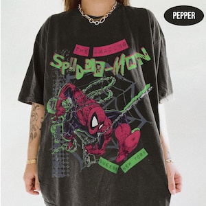 Op de afbeelding: Een antracietgrijs T-shirt met korte mouwen en een afbeelding van het stripfiguur Spider-Man in rood en zwart. De tekst luidt THE AMAZING SPIDER-MAN en QUEENS NEW YORK in het groen. Een zwarte ovaal in de hoek bevat het woord PEPPER.