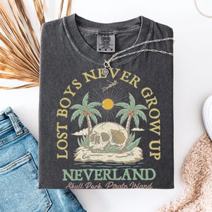May include: Dark grey t-shirt with a graphic featuring a skull, palm trees, and the text "Lost Boys Never Grow Up Neverland Skull Rock Pirate Island." The shirt is laid flat with blue jeans, white shoes, and a necklace.