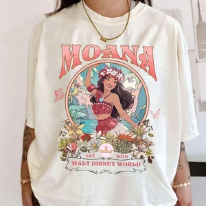 May include: Cream-colored t-shirt featuring a graphic of a woman in a floral crown, with the word "MOANA" in pink above her. The design includes flowers, butterflies, and the text "WALT DISNEY WORLD EST 2016".