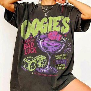 Könnte beinhalten: Ein anthrazitfarbenes Kurzarm-T-Shirt mit einem Grafikdruck mit dem Text OOGIE'S LAIR OF BAD LUCK, WHERE THE ODDS ARE NEVER IN YOUR FAVOR und LET THE BOOGIE TIMES ROLL. Das Design zeigt ein violettes Cocktailglas und zwei Spielwürfel.