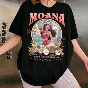 May include: Black t-shirt featuring a colorful graphic of Moana from Walt Disney World. The design includes the word "MOANA" in pink, a floral wreath, and the text "EST 2016". The shirt is a crew neck and has short sleeves.