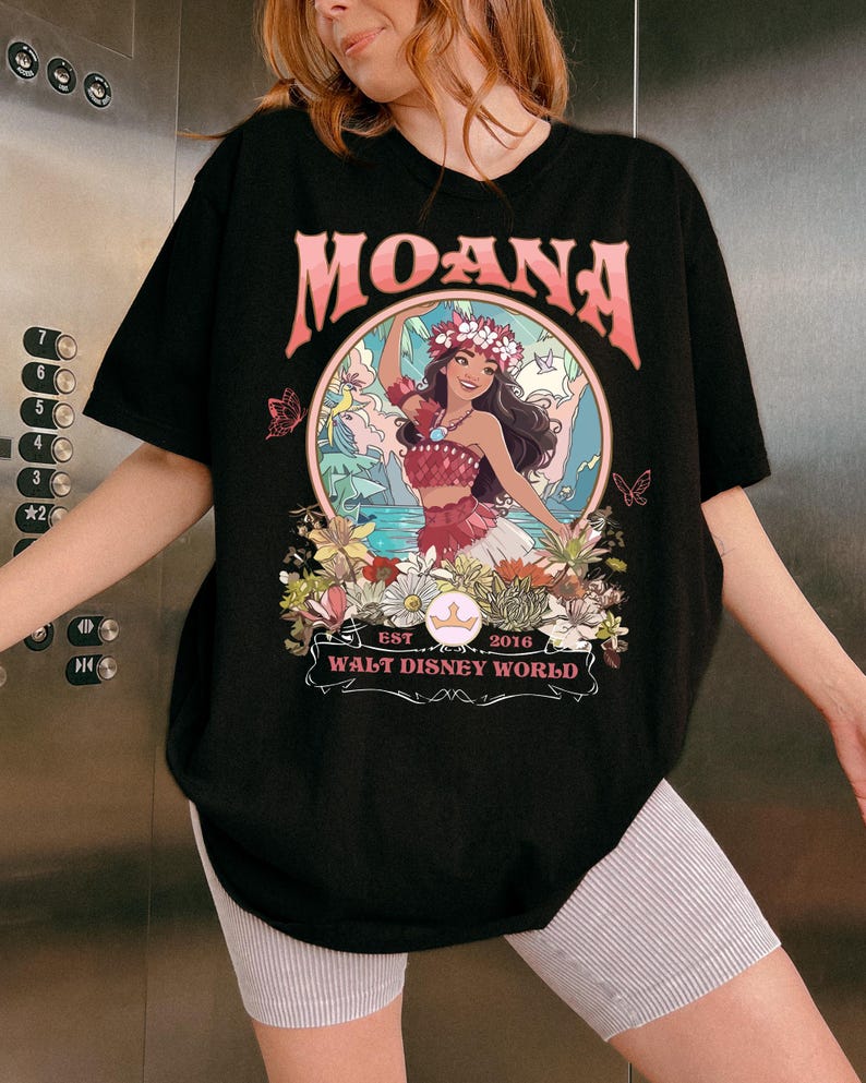 May include: Black t-shirt featuring a colorful graphic of Moana from Walt Disney World. The design includes the word "MOANA" in pink, a floral wreath, and the text "EST 2016". The shirt is a crew neck and has short sleeves.
