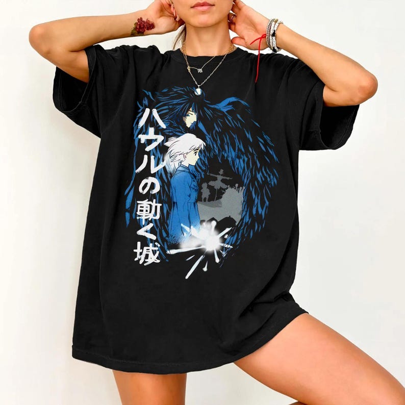 Puede incluir: Camiseta negra con un gr&aacute;fico de la pel&iacute;cula de animaci&oacute;n "El castillo ambulante". El dise&ntilde;o incluye personajes y texto japon&eacute;s. La obra de arte utiliza tonos de azul, blanco y negro, contrastando con el fondo oscuro de la camiseta.