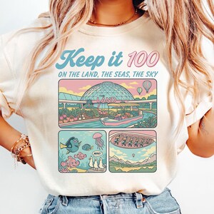 May include: Cream-coloured t-shirt with the text "Keep it 100" in blue and pink. The shirt features a colourful graphic with scenes of a park, underwater life, and hot air balloons. The graphic includes the text "ON THE LAND, THE SEAS, THE SKY" and "THE LAND".