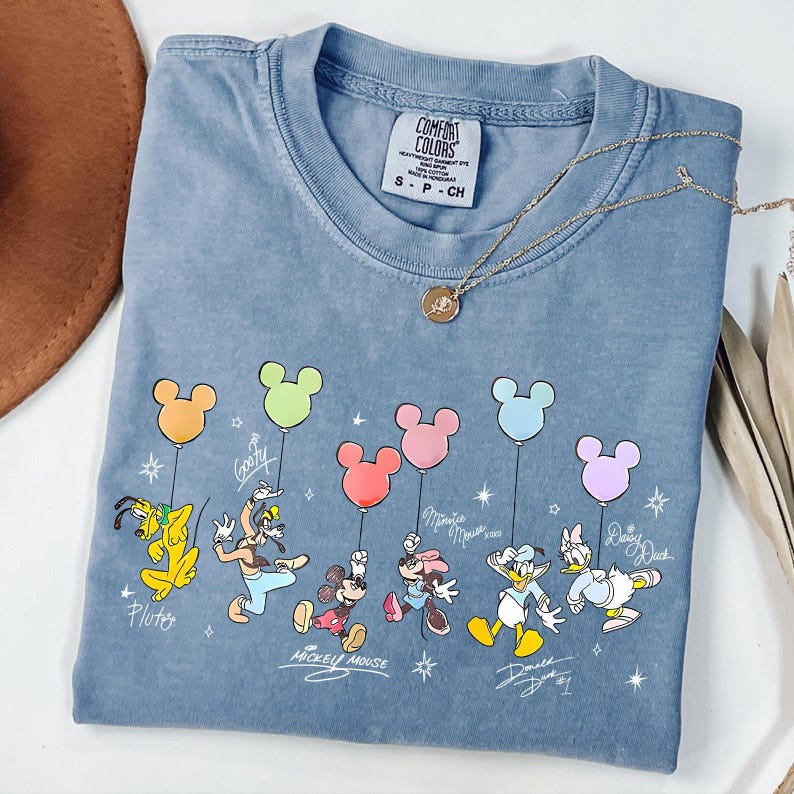 May include: A blue Comfort Colors t-shirt featuring a graphic of Disney characters holding Mickey Mouse-shaped balloons. The characters include Pluto, Goofy, Mickey Mouse, Minnie Mouse, Donald Duck, and Daisy Duck. A gold necklace is draped across the shirt.