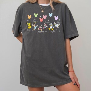 May include: Dark gray t-shirt featuring a colorful graphic of Disney characters holding Mickey Mouse-shaped balloons. The design includes Pluto, Mickey, Minnie, Donald, and Daisy Duck. The shirt has the text "Happy" and character signatures.