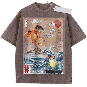 May include: A brown short-sleeve t-shirt featuring a rectangular graphic print of the orange Charizard and blue Blastoise characters. The artwork includes Japanese text, stylized clouds, a red sun, a mountain, and ocean waves in a woodblock style.