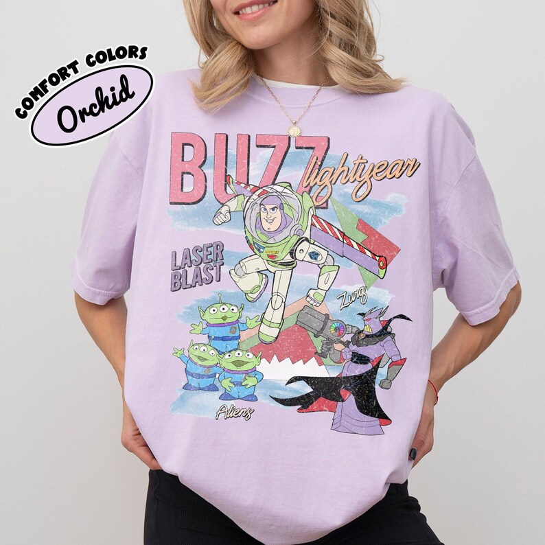 May include: A light purple t-shirt featuring a graphic of Buzz Lightyear and other characters from the movie Toy Story. The design includes the words "Buzz Lightyear" and "Laser Blast" in a retro style. The shirt is labeled "Comfort Colors" and the color is "Orchid".