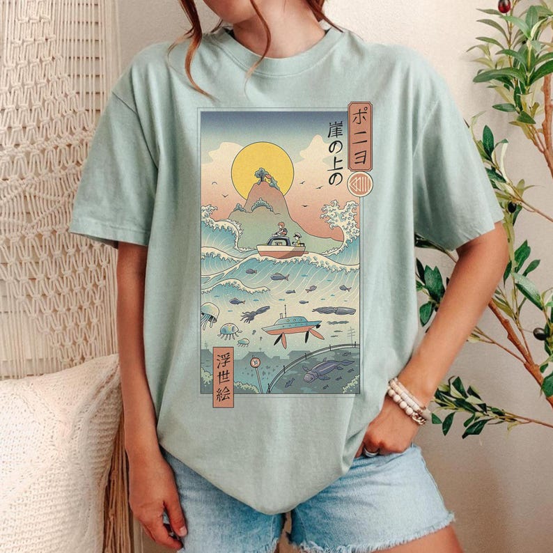 May include: A light green t-shirt featuring a Japanese-style illustration of a boat on ocean waves, a rising sun, and marine life. The artwork includes Japanese text and is printed on the front of the shirt.