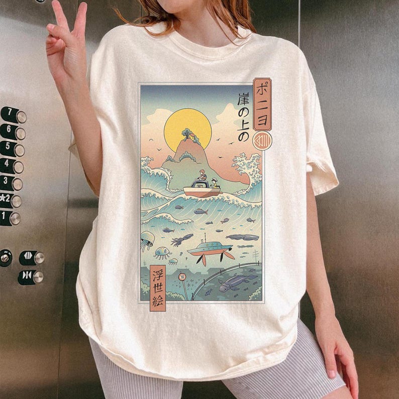 May include: Cream-colored t-shirt featuring a Japanese-inspired graphic. The graphic depicts a boat on ocean waves, a submarine, and a rising sun. Japanese text is visible on the right side of the graphic.