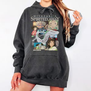 May include: Dark gray hoodie featuring a graphic from the animated film "Spirited Away." The design includes the film's title and various character illustrations. The hoodie has a drawstring hood and a front pocket.
