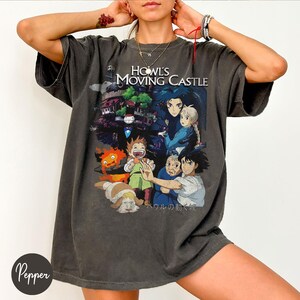 May include: Dark gray t-shirt featuring a colorful graphic from "Howl's Moving Castle." The design includes characters and the film's title. The shirt has short sleeves and a relaxed fit, suitable for casual wear.