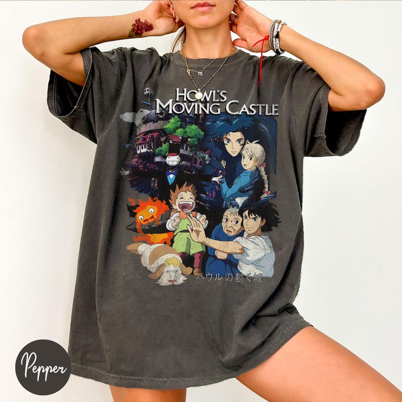 May include: Dark gray t-shirt featuring a colorful graphic from "Howl's Moving Castle." The design includes characters and the film's title. The shirt has short sleeves and a relaxed fit, suitable for casual wear.