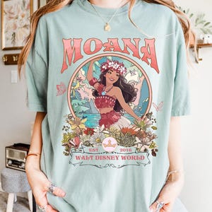 May include: A light blue t-shirt featuring a graphic of Moana with the text "MOANA" in a coral color. The graphic includes a floral design and the text "EST 2016 WALT DISNEY WORLD".