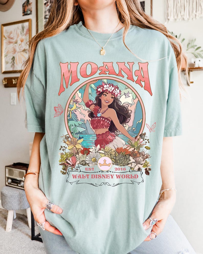 May include: A light blue t-shirt featuring a graphic of Moana with the text "MOANA" in a coral color. The graphic includes a floral design and the text "EST 2016 WALT DISNEY WORLD".
