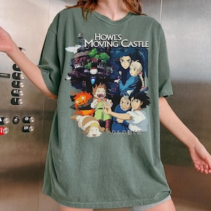 May include: Sage green t-shirt featuring a colorful graphic from "Howl's Moving Castle." The design includes characters and the film's title. The shirt has short sleeves and a crew neck.