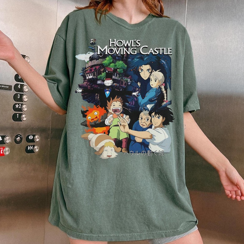 May include: Sage green t-shirt featuring a colorful graphic from "Howl's Moving Castle." The design includes characters and the film's title. The shirt has short sleeves and a crew neck.