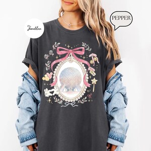 May include: Dark gray t-shirt featuring a watercolor design of a globe within a pink bow-framed oval. The design includes floral elements, Mickey Mouse, and a magic wand. The word "Pepper" is in a speech bubble.
