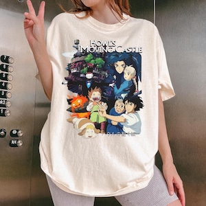 May include: Cream-colored t-shirt featuring a colorful graphic from the animated film "Howl's Moving Castle." The design includes characters and the film's title. The shirt is a relaxed fit.