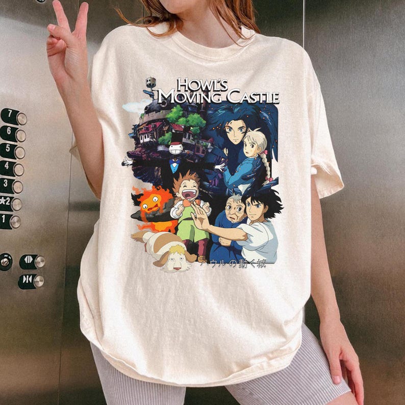 May include: Cream-colored t-shirt featuring a colorful graphic from the animated film "Howl's Moving Castle." The design includes characters and the film's title. The shirt is a relaxed fit.