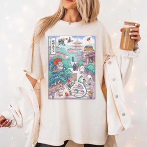 Puede incluir: Camiseta color crema con una colorida ilustraci&oacute;n de estilo anime. La obra de arte representa una escena vibrante con personajes, edificios y un drag&oacute;n sinuoso. El texto japon&eacute;s es visible en el lado izquierdo del gr&aacute;fico. La camiseta tiene un corte holgado.