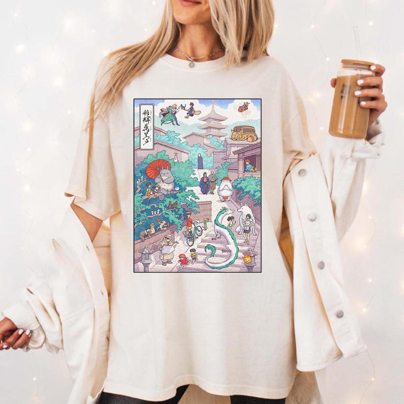 Puede incluir: Camiseta color crema con una colorida ilustraci&oacute;n de estilo anime. La obra de arte representa una escena vibrante con personajes, edificios y un drag&oacute;n sinuoso. El texto japon&eacute;s es visible en el lado izquierdo del gr&aacute;fico. La camiseta tiene un corte holgado.