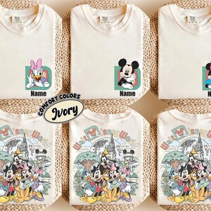 Könnte beinhalten: Sechs elfenbeinfarbene T-Shirts mit Disney-Charaktergrafiken. Die obere Reihe zeigt einzelne Charaktere mit einem Buchstaben und einem Namensfeld. Die untere Reihe zeigt eine Gruppenillustration von Disney-Charakteren mit dem Text Walt Disney World.