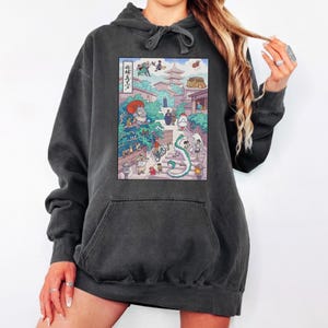 Puede incluir: Sudadera con capucha gris oscuro con una colorida ilustraci&oacute;n de una escena de inspiraci&oacute;n japonesa, con un drag&oacute;n, personajes y edificios. La obra de arte est&aacute; centrada en la parte delantera, con un bolsillo de canguro y una capucha con cord&oacute;n.