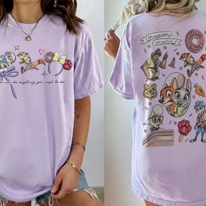 Puede incluir: Camiseta lila con personajes de dibujos animados y texto de Zootopia. La parte delantera muestra a Judy Hopps, Nick Wilde y accesorios tem&aacute;ticos. La parte trasera presenta m&aacute;s personajes y la frase "The Big Donut".
