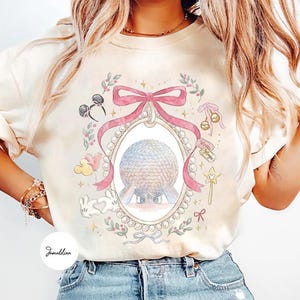 May include: Cream-colored t-shirt featuring a watercolor design with an oval frame, a pink bow, and a sphere. The design includes Mickey Mouse ears, balloons, a rabbit, and a magic wand. The words "Magic" are visible on a tag.