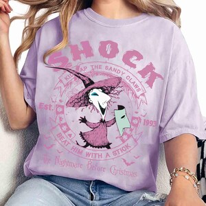 Op de afbeelding: Een lichtpaars t-shirt met een afbeelding van het personage Shock uit The Nightmare Before Christmas. Het ontwerp bevat de tekst SHOCK, KIDNAP THE SANDY CLAWS, BEAT HIM WITH A STICK, Est. 1993 en The Nightmare Before Christmas.