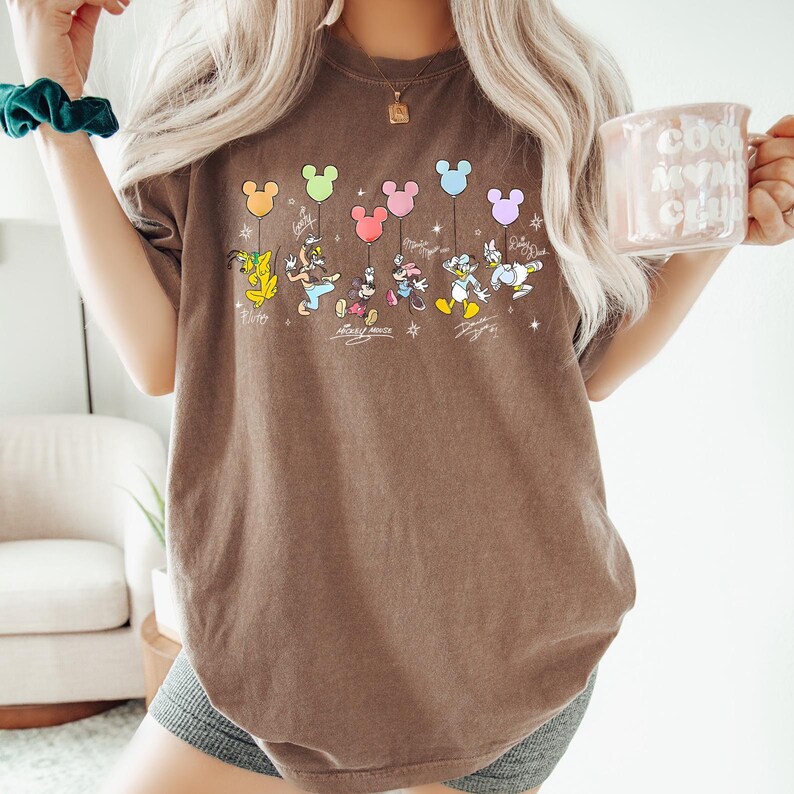 May include: Brown t-shirt featuring a colorful graphic of Disney characters holding Mickey Mouse-shaped balloons. The characters include Goofy, Donald Duck, Mickey Mouse, Minnie Mouse, Daisy Duck, and Pluto. Each character has a signature below them.