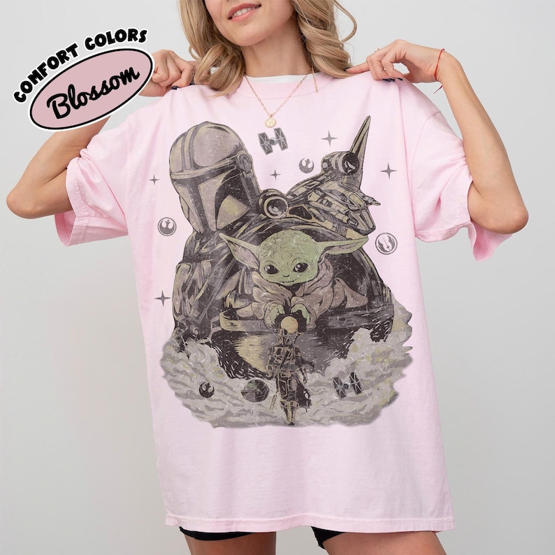 Może przedstawiać: Jasnor&oacute;żowy t-shirt z nadrukiem graficznym przedstawiającym postać w hełmie, małe zielone stworzenie oraz statek kosmiczny. Wz&oacute;r zawiera ikony gwiazd i okrągłe symbole. Tekst Comfort Colors Blossom znajduje się w lewym g&oacute;rnym rogu.