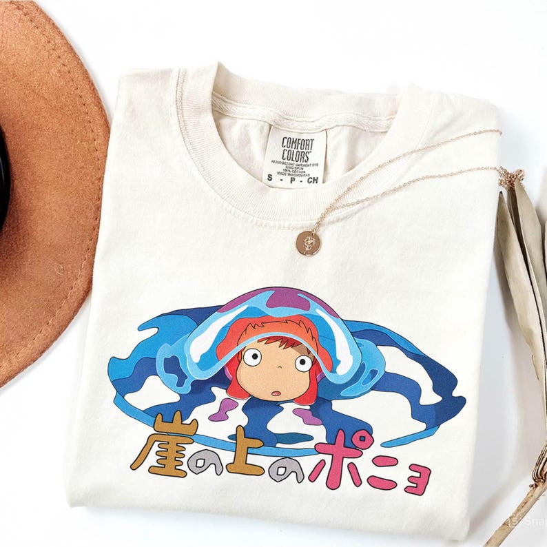 May include: Cream-colored t-shirt featuring a colorful graphic of a cartoon character with red hair and a blue and purple design. The Japanese text is below the character. A gold necklace and a brown hat are also in the image.