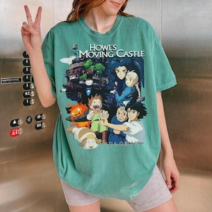 May include: A teal-colored t-shirt featuring a graphic from the animated film "Howl's Moving Castle." The design includes characters and the film's title. The shirt is worn by a person.